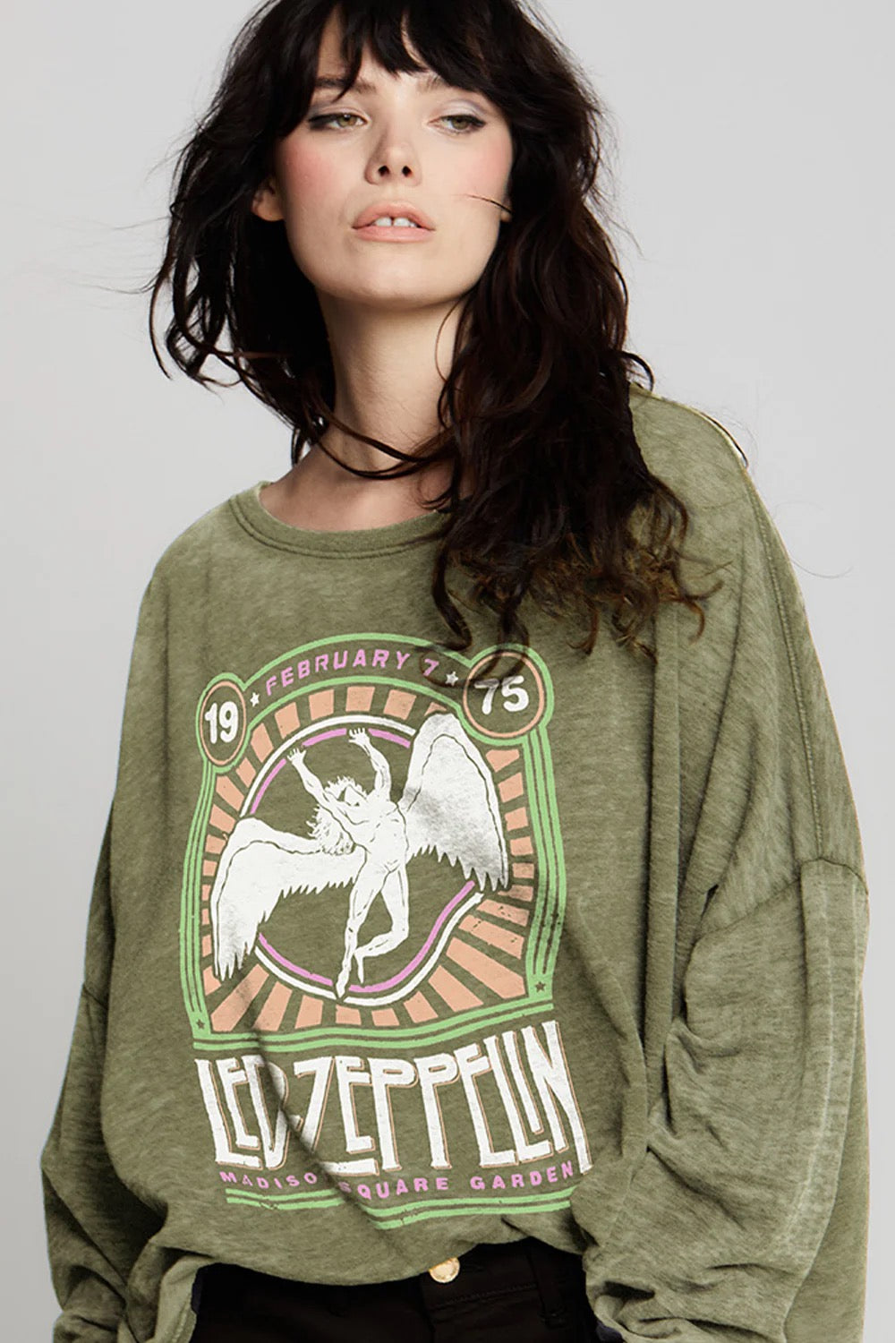 A woman with wavy dark hair wears the Recycled Karma Led Zeppelin Msg Ls—a vintage-inspired, oversized olive sweatshirt featuring an angel graphic and referencing Madison Square Garden, 1975. Its relaxed, oversized fit adds effortless style as she gazes off to the side against a plain background.