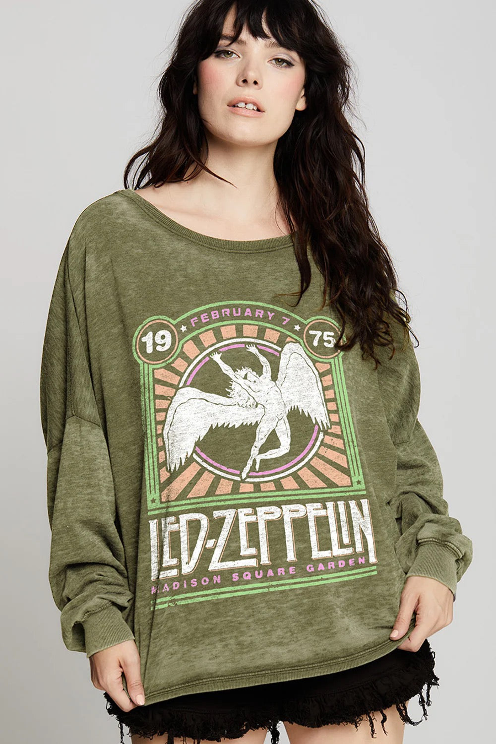 A woman with wavy dark hair wears the Recycled Karma Led Zeppelin Msg Ls in olive, its vintage-inspired oversized fit styled with black shorts, standing against a neutral background.