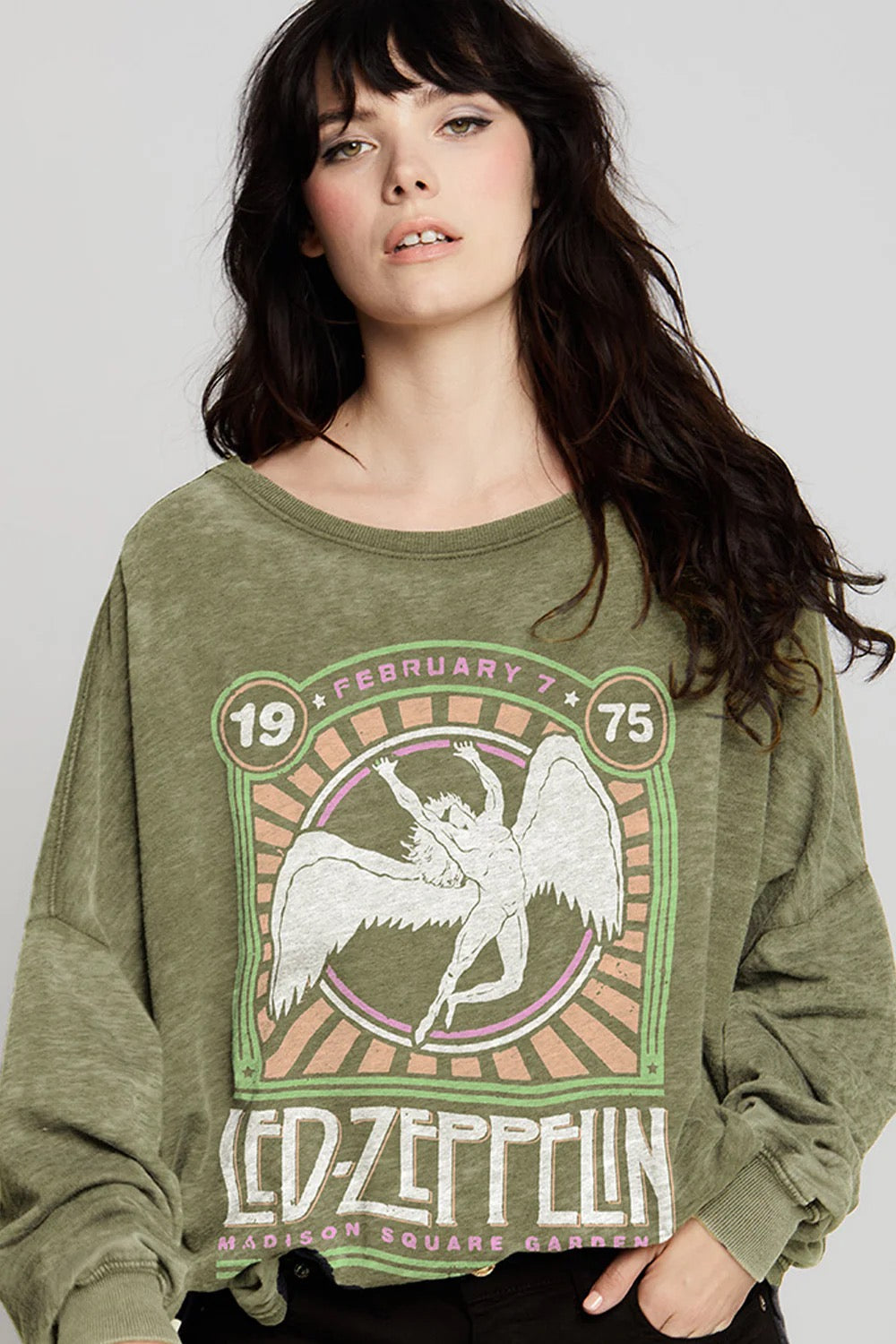 A woman with long dark hair wears the Recycled Karma Led Zeppelin Msg Ls, a vintage-inspired oversized green sweatshirt featuring a winged figure and “February 7, 1975.” Its relaxed drape adds effortless style as she stands against a plain light background, gazing slightly upward.
