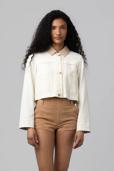 A woman with long curly hair wears the Morning Zen Jacket by Blank in cream, featuring large front pockets and long sleeves, paired with high-waisted tan shorts. The cropped silhouette stands out against a plain gray background.