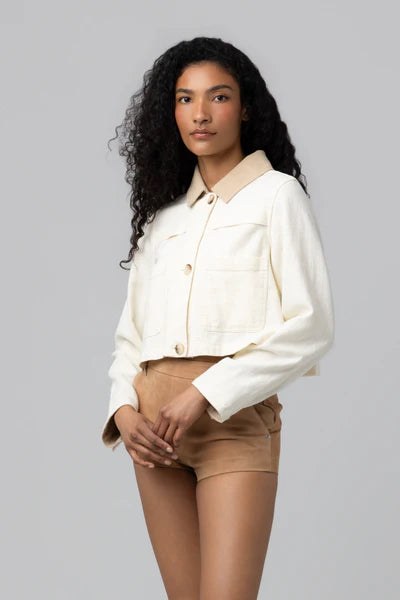 A woman with long curly hair stands against a plain background, wearing Blank's Morning Zen Jacket—a cream-colored cropped piece with large front pockets and beige collar—paired with tan high-waisted shorts in soft neutral tones.