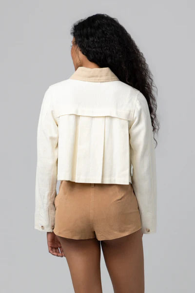 A woman with long, curly hair stands with her back to the camera, wearing the Morning Zen Jacket by Blank—a cropped cream silhouette—paired with high-waisted tan shorts against a plain gray background.