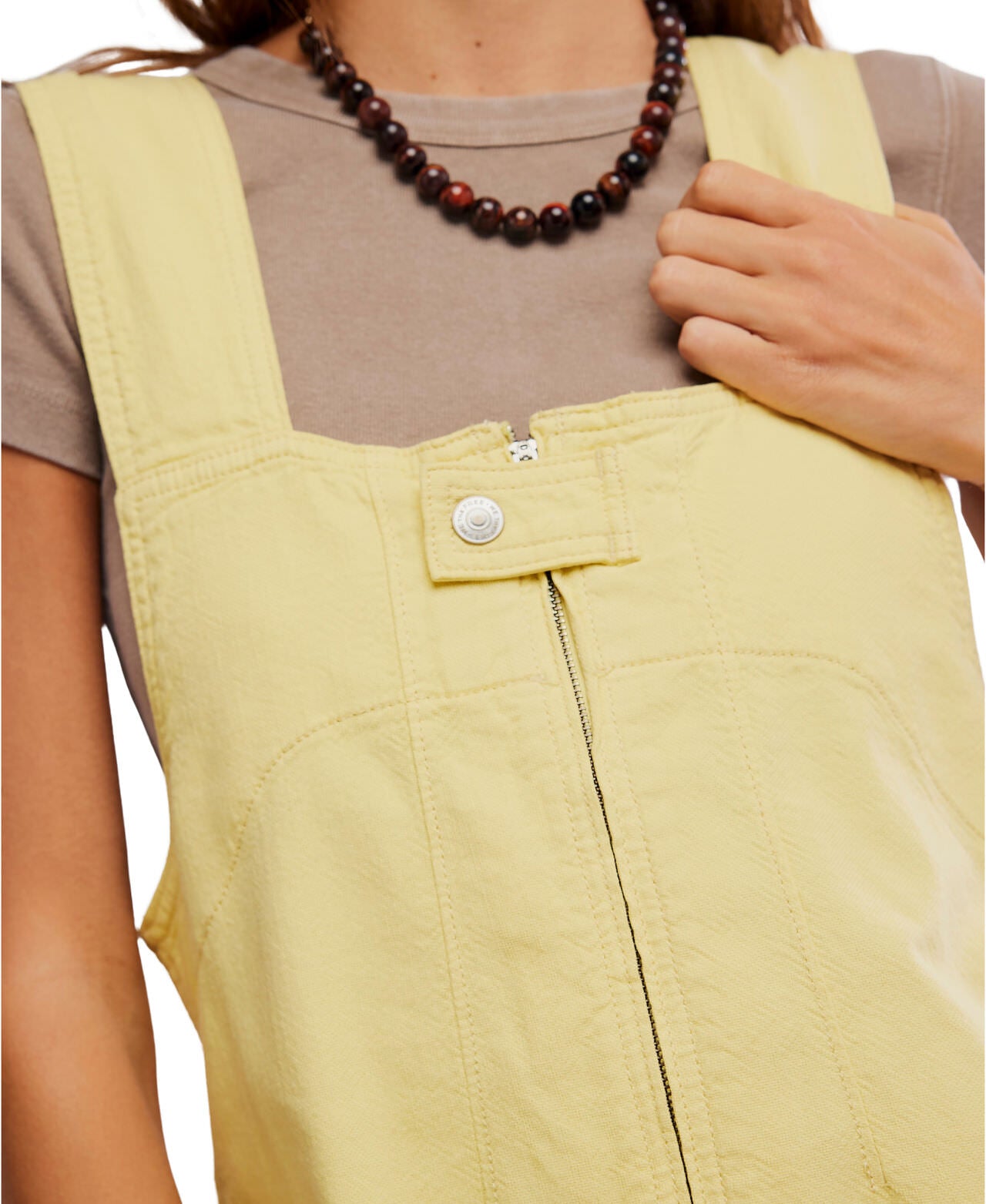 A person wears Free People's Jude Shortall—a sleeveless, light yellow zip-up dress with a top button—over a beige T-shirt, accessorized with a brown beaded necklace.