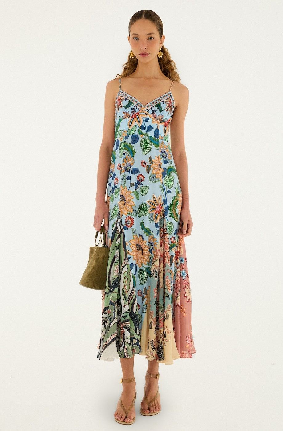 A woman faces forward in the Farm Rio Floral Delhi Light Blue Sleeveless Maxi dress, which features a colorful floral print and asymmetrical hem. She carries an olive-green handbag, wears strappy sandals, and has loose waves in her hair.