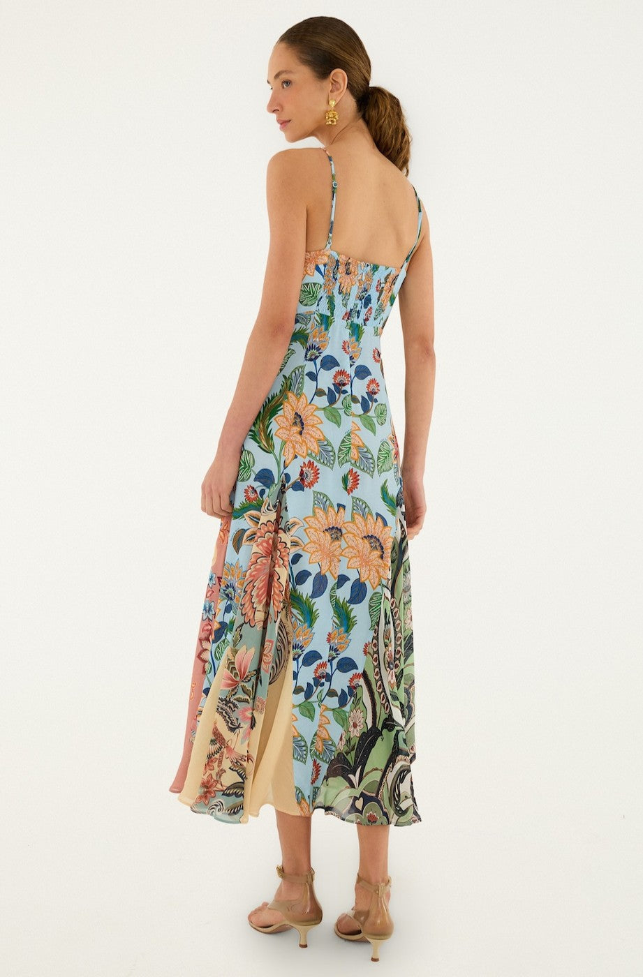 A woman wears the Farm Rio Floral Delhi Light Blue Sleeveless Maxi, featuring delicate florals and a flared hem. She pairs it with dangling earrings, nude heels, and has her hair in a low ponytail, turned slightly to the side.