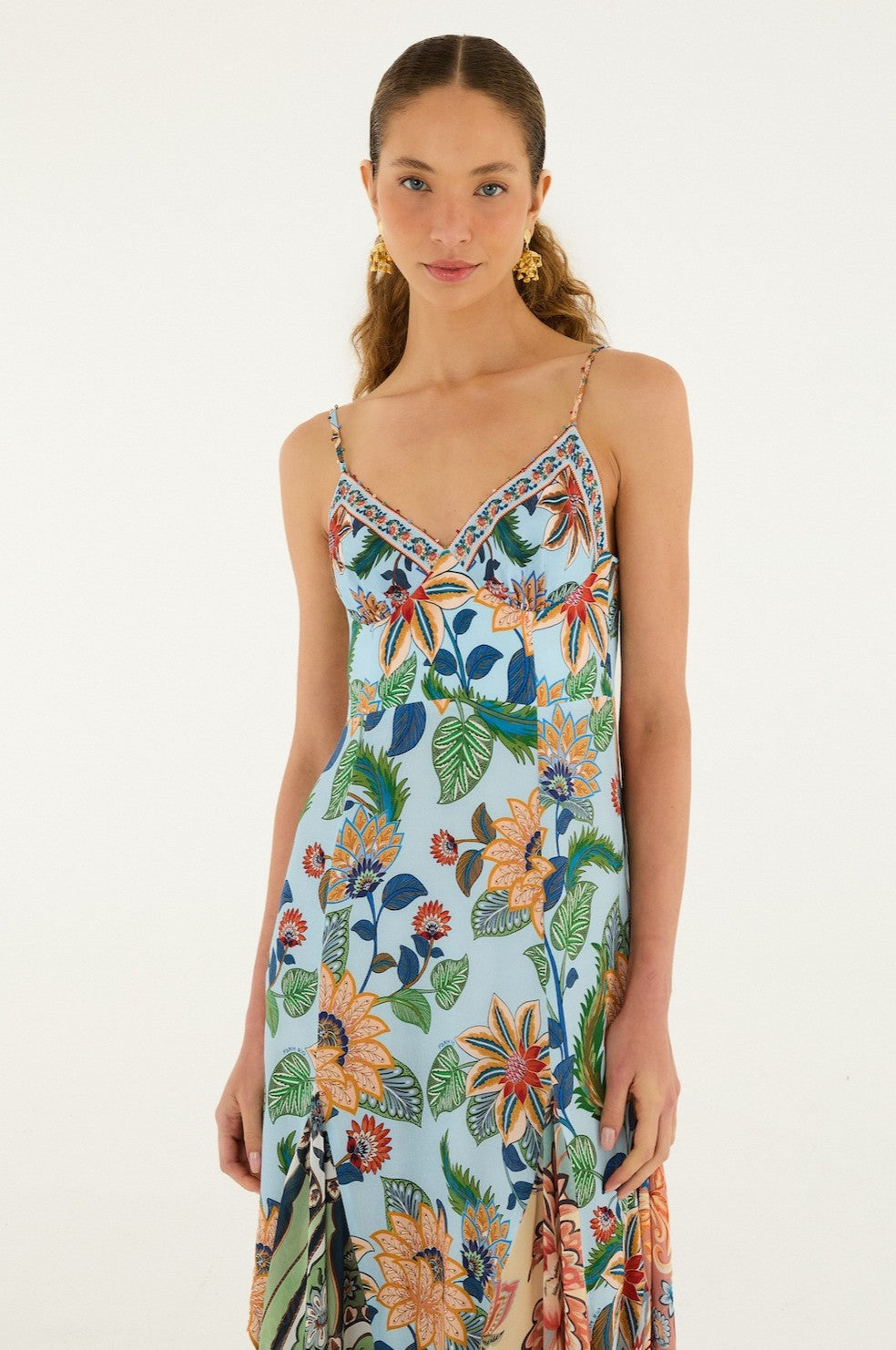A woman models the Farm Rio Floral Delhi Light Blue Sleeveless Maxi, featuring floral patterns, thin straps, and a flowing silhouette. She stands against a white background, wearing gold earrings with her long brown hair pulled back.