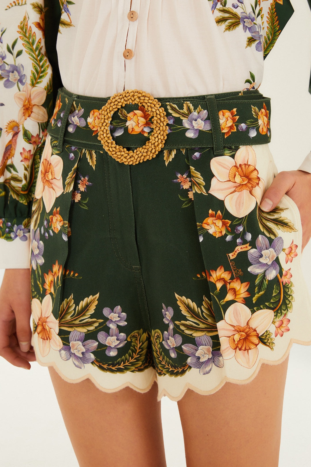A person wears Farm Rio's Berry Garden Twill Shorts in Black Multi, featuring a woven buckle belt, colorful floral prints, and cream scalloped hems; part of a floral blouse is also visible.