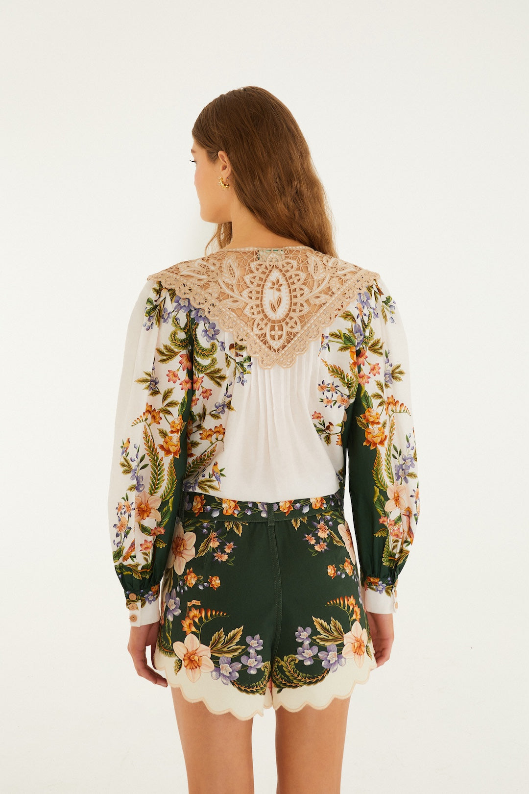 A woman with long brown hair faces away, pairing a white lace floral blouse with Farm Rio's Berry Garden Twill Shorts, featuring colorful flowers, high waist, and a chic buckle belt.