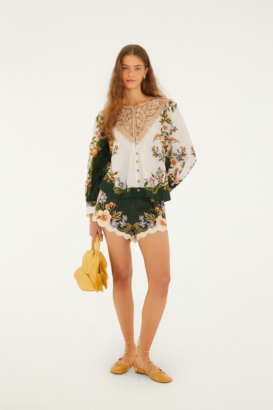 A woman with long, wavy hair and a neutral expression stands against a plain background wearing Farm Rio's Berry Garden Twill Shorts, a floral embroidered blouse with lace detail, yellow sandals, and holding a yellow handbag.
