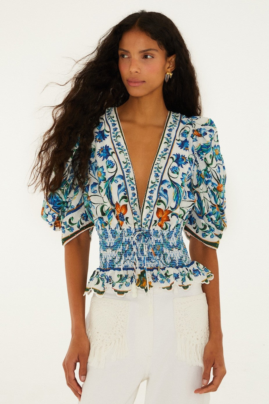 A woman with long wavy hair wears the Farm Rio Flora Tile Off White Top in Blue Multi, featuring puffed sleeves, a deep V-neck, and a smocked waist, paired with textured white pants. She stands against a plain white background.