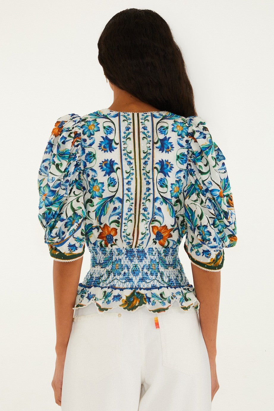 A woman with long dark hair is seen from the back wearing the Farm Rio Flora Tile Off White Top, an organic cotton blouse with a colorful floral tile print and puffed sleeves, paired with white pants against a light background.