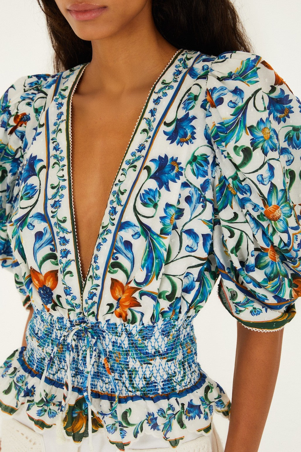 A woman wears the Farm Rio Flora Tile Off White Top, featuring organic cotton, a deep V-neck, puffed sleeves, and a vibrant blue, green, and orange floral tile print. The smocked bodice gently cinches her waist; her face is partially out of frame.