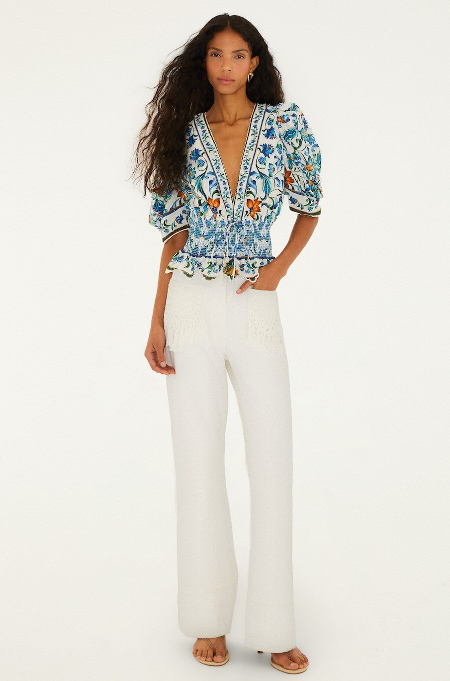 A woman with long wavy hair wears the Farm Rio Flora Tile Off White Top with puffed sleeves and a floral tile pattern, paired with high-waisted, wide-leg white trousers. She stands barefoot against a plain white background.