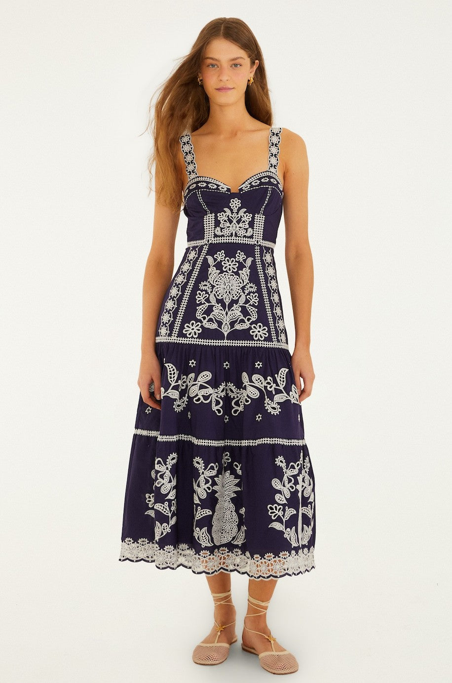 A woman with long brown hair wears the Blue And White Richillieu Midi Dress by Farm Rio in Blue Multi, featuring floral embroidery and lace accents. She stands facing forward with a neutral expression, wearing beige lace-up flats.