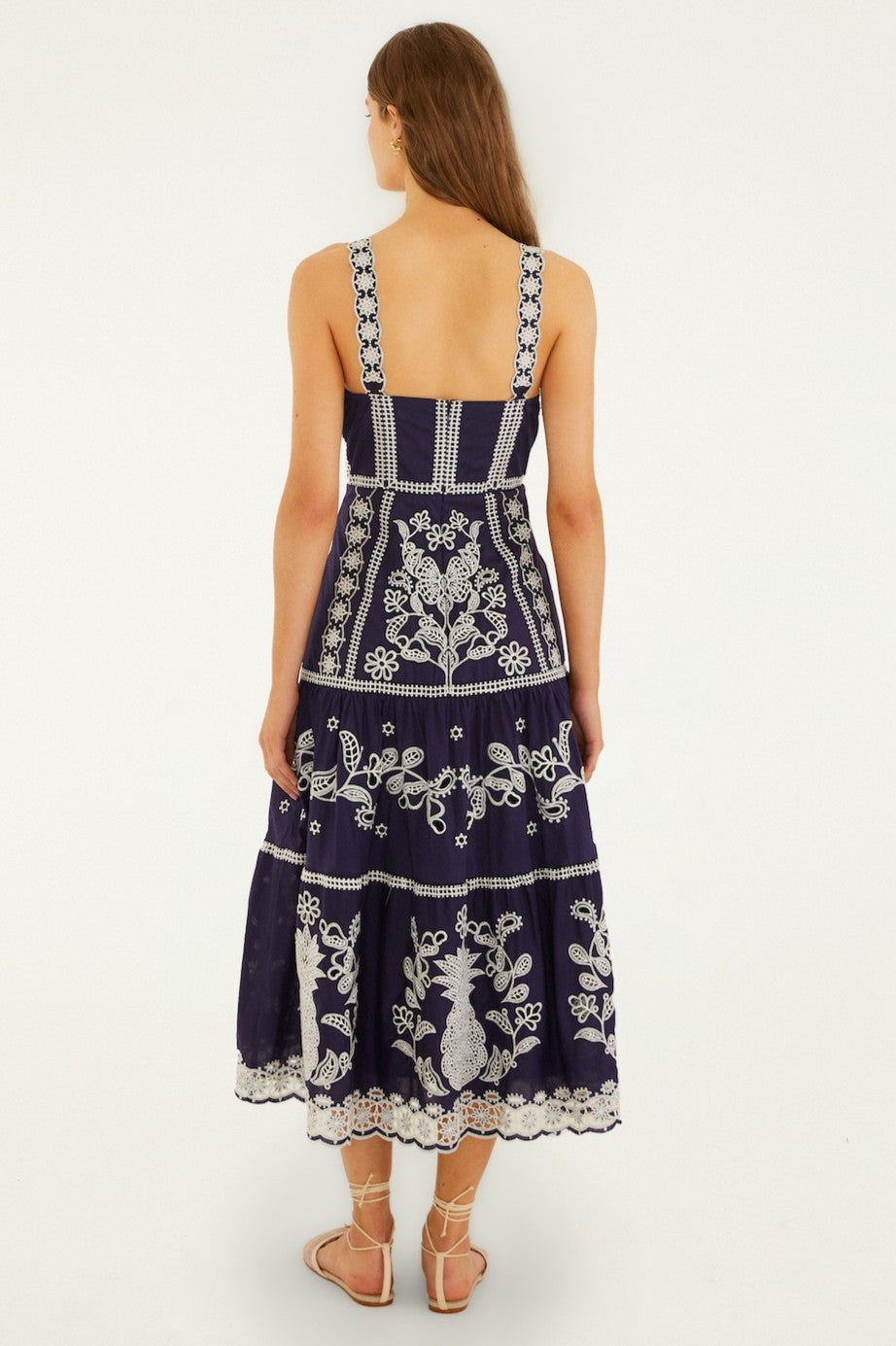 A woman with long brown hair stands facing away, wearing the Farm Rio Blue And White Richillieu Midi Dress. This 100% cotton dress features white embroidery, lace details, a square back, and a midi silhouette for effortless Brazilian style.