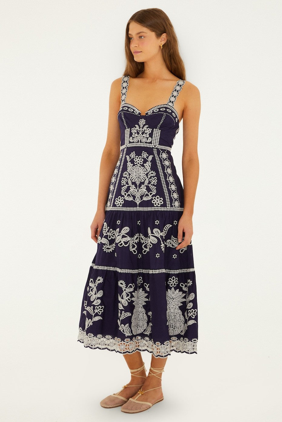 A woman wears the Farm Rio Blue And White Richillieu Midi Dress, a navy sleeveless 100% cotton piece with white embroidered floral patterns, paired with beige lace-up flats. She has long brown hair and looks to the side against a plain background.