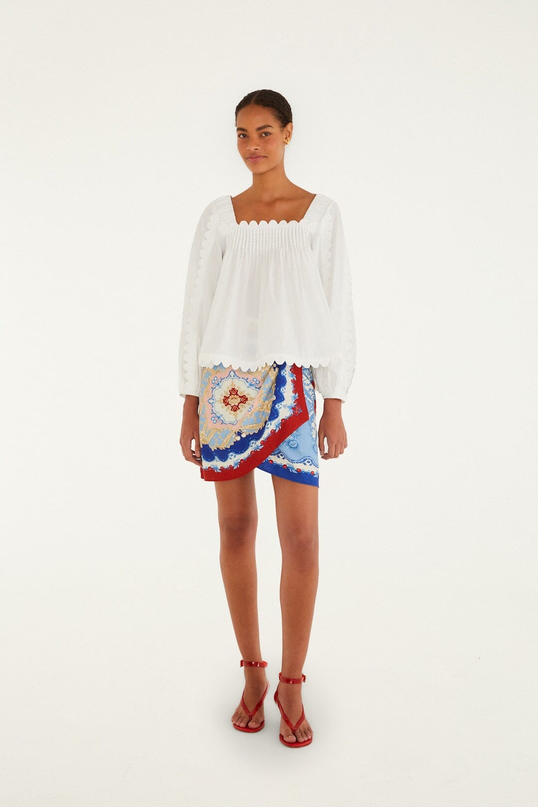 A woman poses against a white backdrop in a white long-sleeve, square-neck blouse, red strappy sandals, and the Farm Rio Mixed Cherry Royalty Scarf Mini Skirt featuring a vibrant blue, red, and white print.