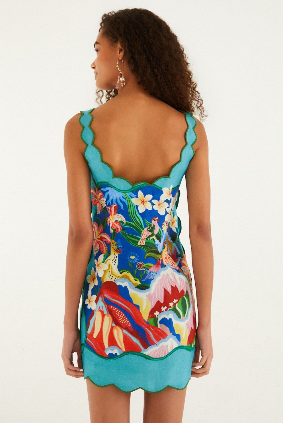 A woman with curly hair faces away, wearing the Farm Rio Garden Of Life Scarf Multicolor Sleeveless dress featuring a square neck, scalloped edges, and a vibrant tropical print. The background is plain white.