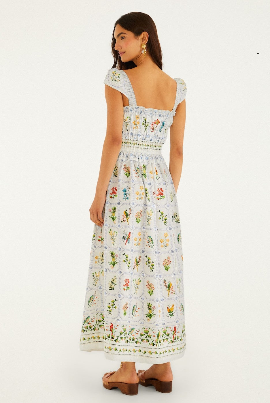 A woman with long dark hair stands facing away, wearing the Farm Rio Tropical Porcelain Off-White Maxi Dress featuring a smocked bodice, puff sleeves, and a vibrant botanical print. She pairs it with sandals and earrings.