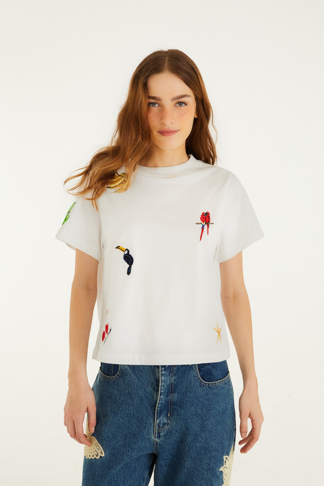 A woman with long, light brown hair wears the Farm Rio Cool Day Relaxed T-shirt in White Multi, featuring colorful embroidered birds, paired with blue jeans with lace patches, standing against a plain white background.