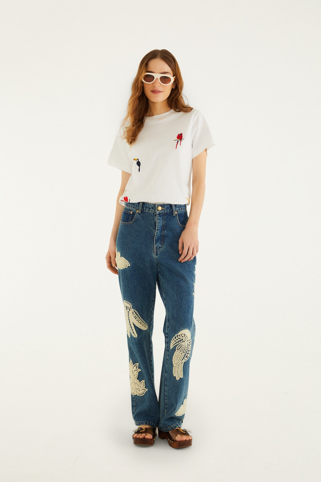 A woman with long hair wears white sunglasses, brown sandals, blue jeans with white embroidery, and the Cool Day Relaxed T-shirt by Farm Rio, featuring small red and blue designs, standing against a plain white background.
