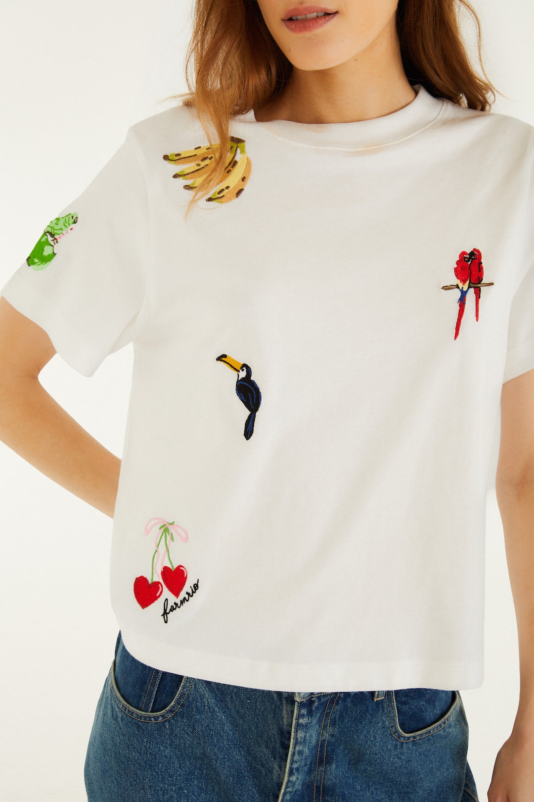 A woman wears the Cool Day Relaxed T-shirt by Farm Rio, an off-white soft cotton top with embroidered bananas, a green bird, a red parrot, a black snake, cherries with "Amour," paired effortlessly with blue jeans.