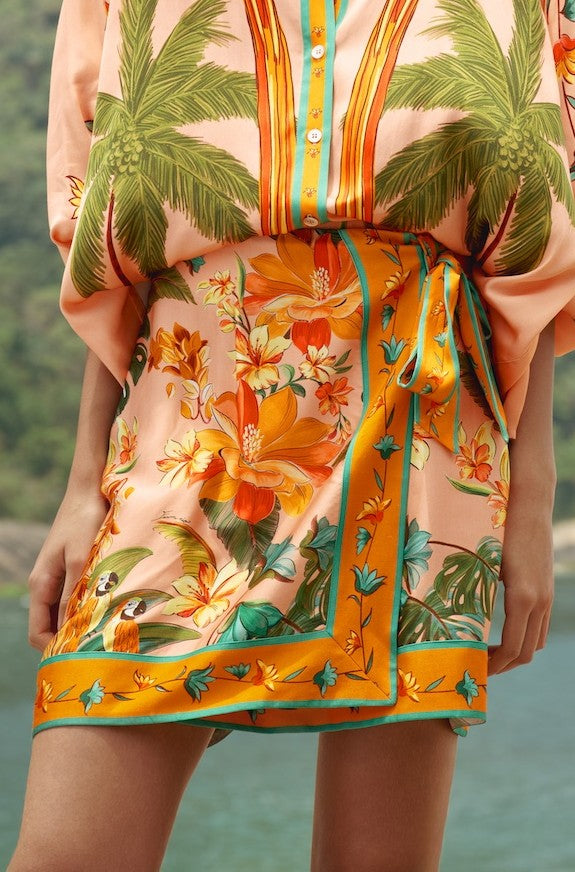 A person by the water wears Farm Rio's Birds Mini Skirt—100% viscose with bright florals and birds—paired with a palm-pattern blouse and styled with a flattering self-tie waist.