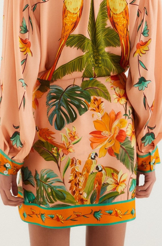 A person wearing the Farm Rio Birds Mini Skirt, featuring a peach base with vibrant tropical parrots, large flowers, and green leaves. The image highlights the self-tie waist at the lower back, plus skirt and sleeves. Made from 100% viscose.