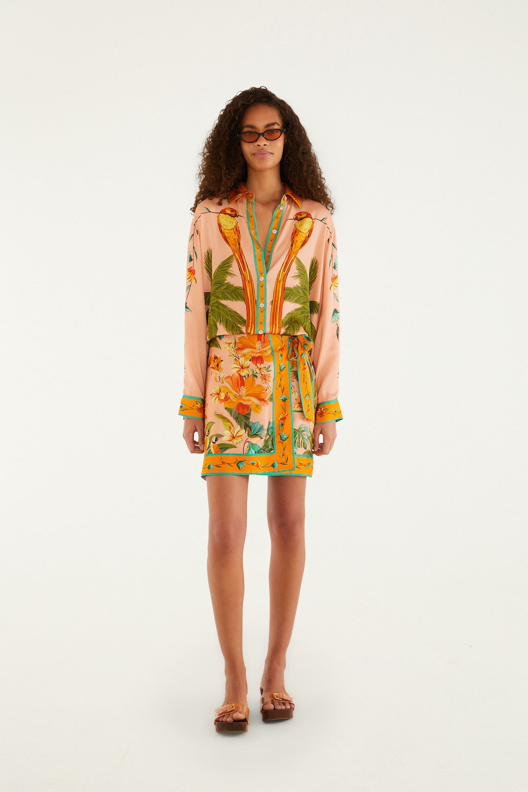 A woman with curly hair and glasses faces forward in the Farm Rio Birds Mini Skirt, featuring tropical floral and palm leaf patterns, an adjustable waist, orange trim, and sandals, set against a plain white background.