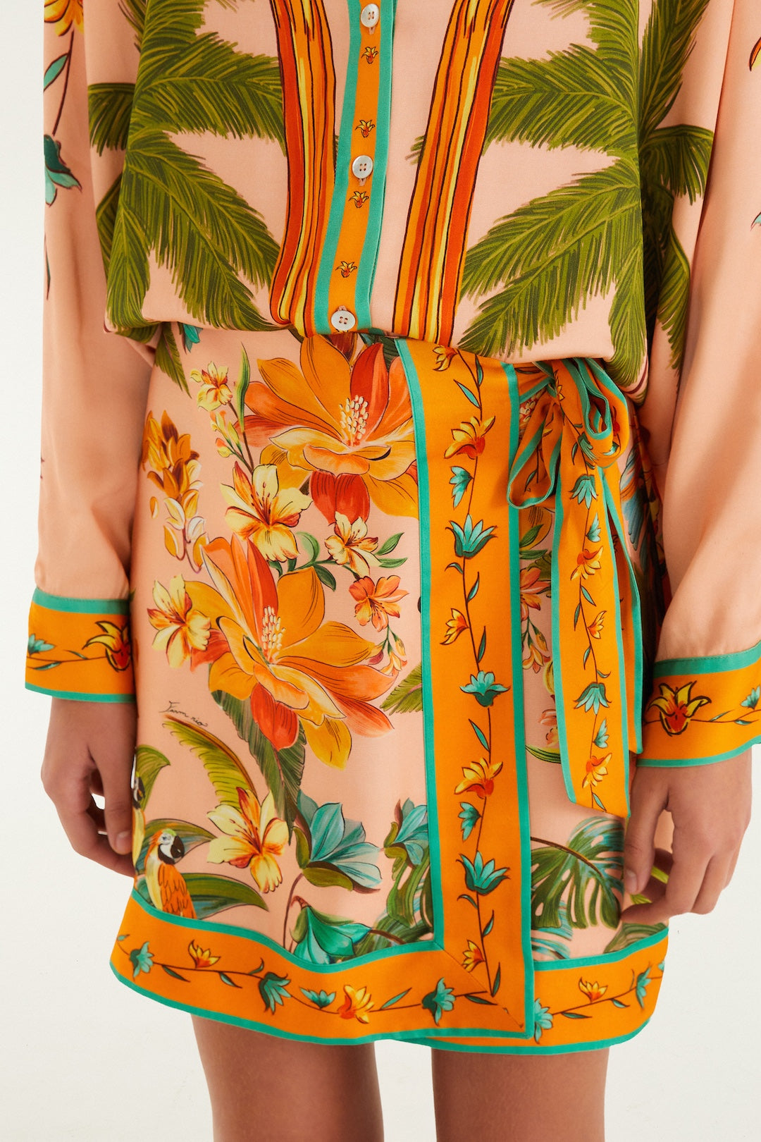 A person wears the Farm Rio Birds Mini Skirt, a tropical-themed piece with palm leaves and orange flowers on peach, featuring a self-tie waist, paired with a matching long-sleeve top accented by turquoise and orange borders.