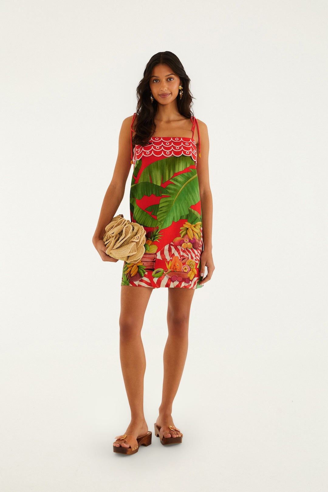 A woman models the Farm Rio Fruit Trolley Red Mini Dress—a vibrant, fruit print mini with spaghetti straps—paired with a tan clutch and brown heeled sandals. She stands against a white background, her long waves complementing her relaxed, friendly vibe.