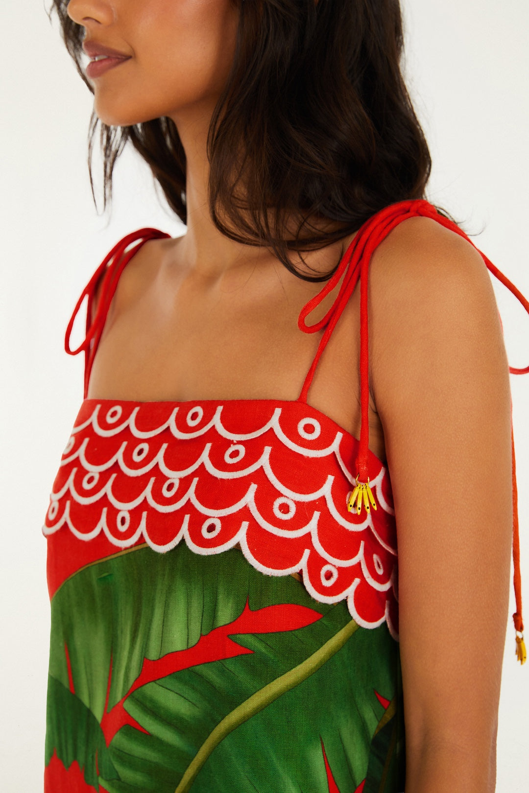 A woman with wavy dark hair wears the Fruit Trolley Red Mini Dress by Farm Rio, featuring a fruit print, red shoulder ties, a red and white scalloped bodice, and large green leaves. The mini silhouette pops against the plain white background.
