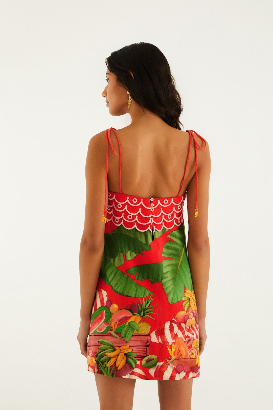 A woman with long dark hair faces away, wearing the Farm Rio Fruit Trolley Red Mini Dress—featuring a vibrant fruit print with green leaves, spaghetti straps, a low back, and a mini silhouette—against a plain white background.