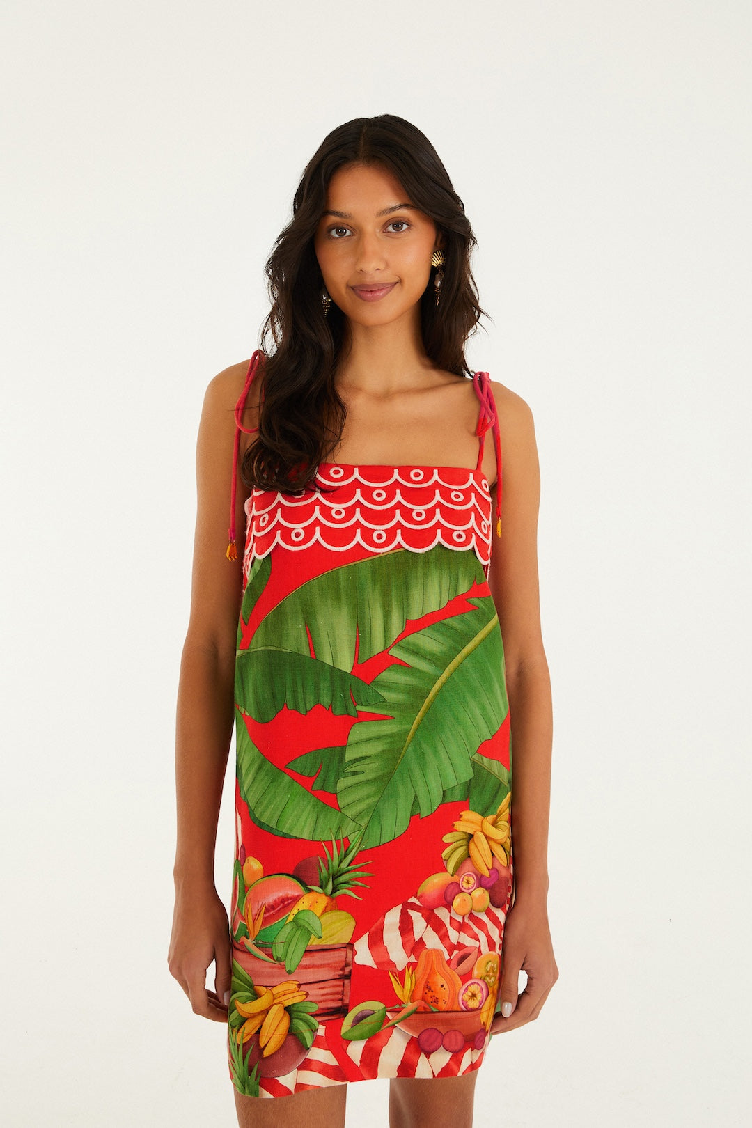 A woman with long dark hair wears the Farm Rio Fruit Trolley Red Mini Dress in Red Multi, featuring a sleeveless design with spaghetti straps and vibrant tropical fruit and green leaf patterns, posed against a plain white background.