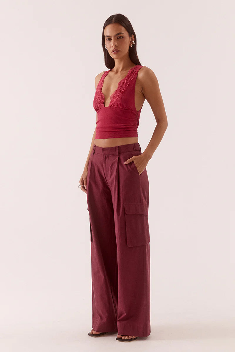 A woman stands against a light background wearing the Sovere Loni Top, a sleeveless deep V-neck style with lace trim, paired with wide-legged maroon cargo pants and her hands in her pockets.