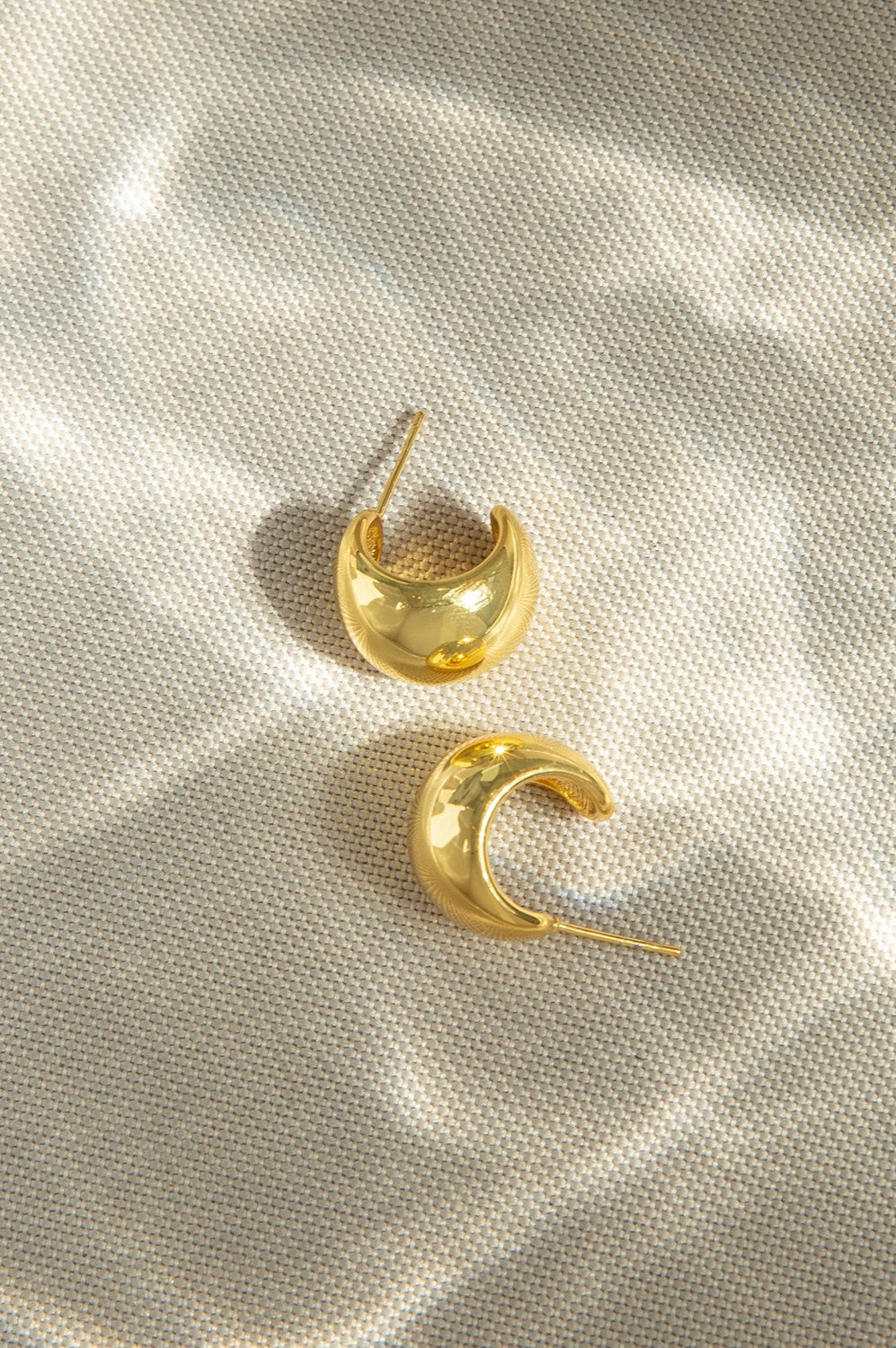 Two gold Waterproof Stainless Chubby Post Hoop earrings by Marlyn Schiff rest on a textured, light-colored fabric, illuminated by soft, dappled sunlight reflections.
