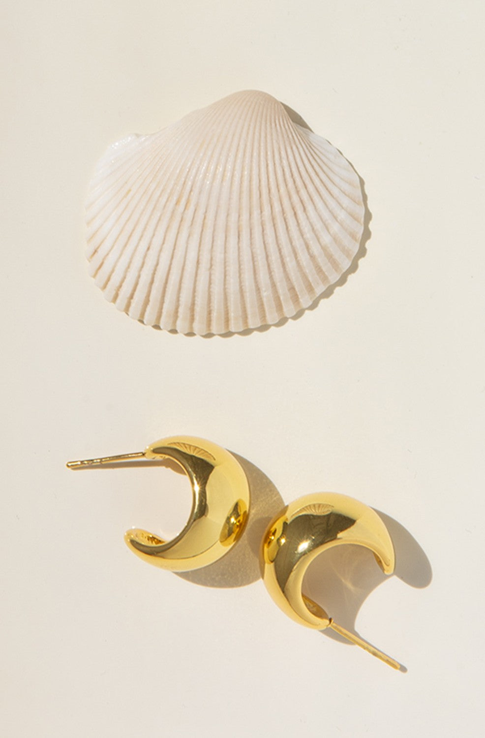 A pair of Marlyn Schiff Waterproof Stainless Chubby Post Hoops, featuring a shiny gold crescent-shaped design and lustrous PVD plating, is displayed with a white seashell on a light beige background.