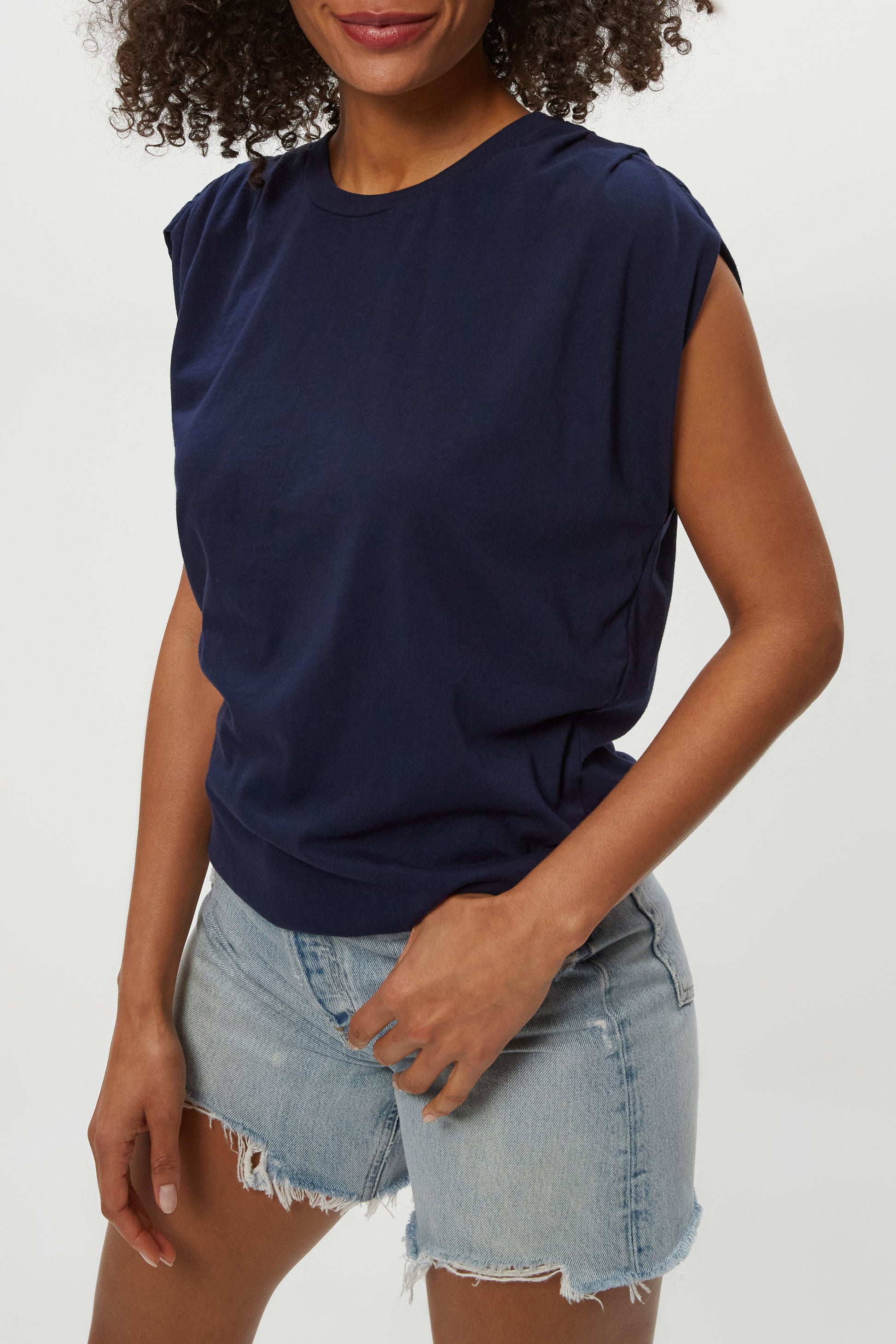 A woman stands against a plain background, smiling slightly with one hand on her hip, wearing the Michael Stars Luca Bubble Tee in navy blue and light blue denim shorts with frayed hems.