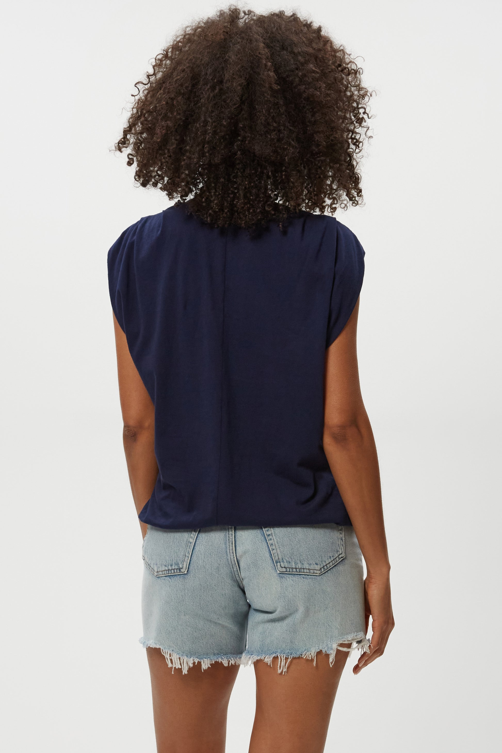 A person with curly hair is shown from behind, wearing the Michael Stars Luca Bubble Tee in navy and light blue frayed denim shorts, styled against a plain white background.