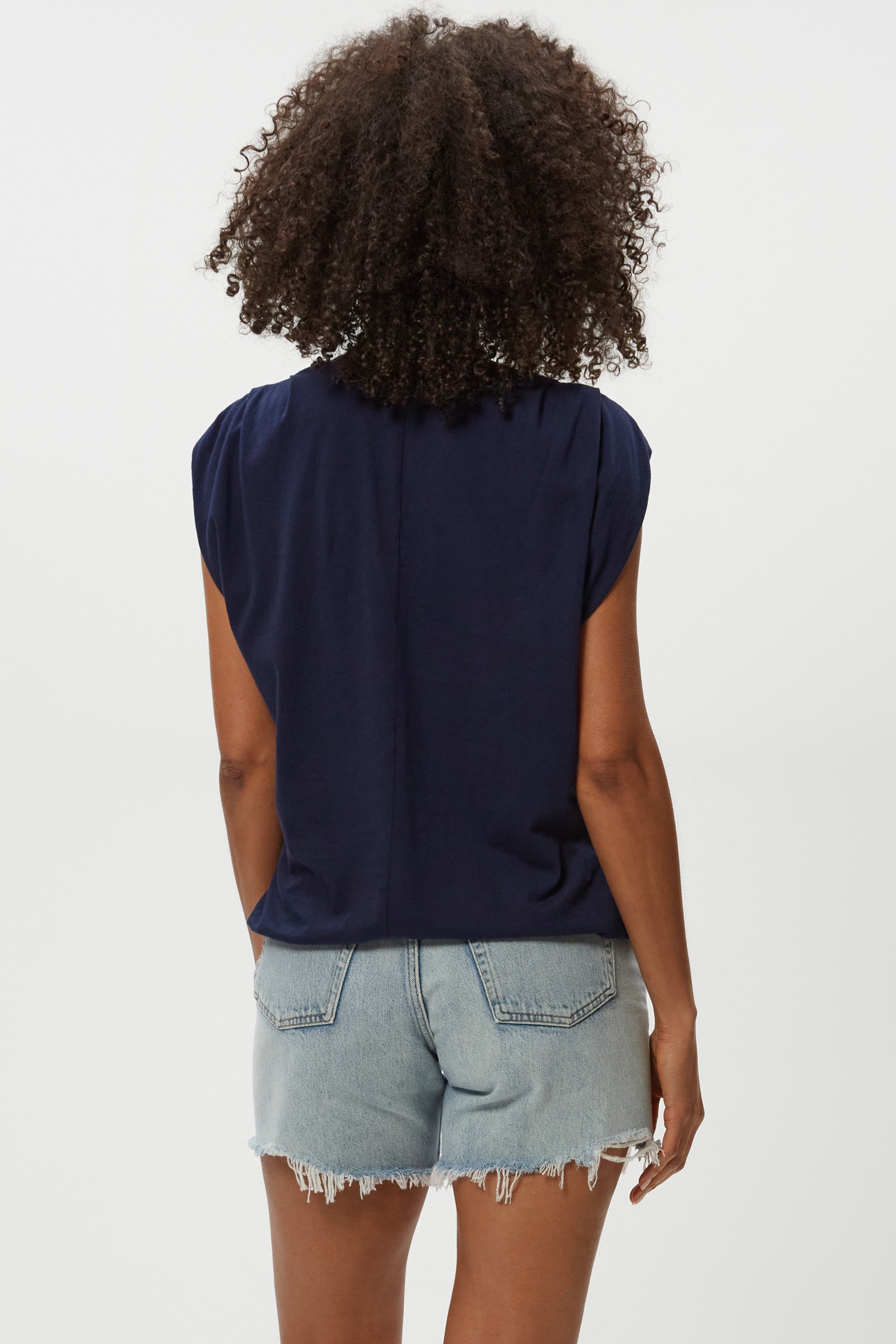 A person with curly hair is shown from behind, wearing the Michael Stars Luca Bubble Tee in navy and light blue frayed denim shorts, styled against a plain white background.