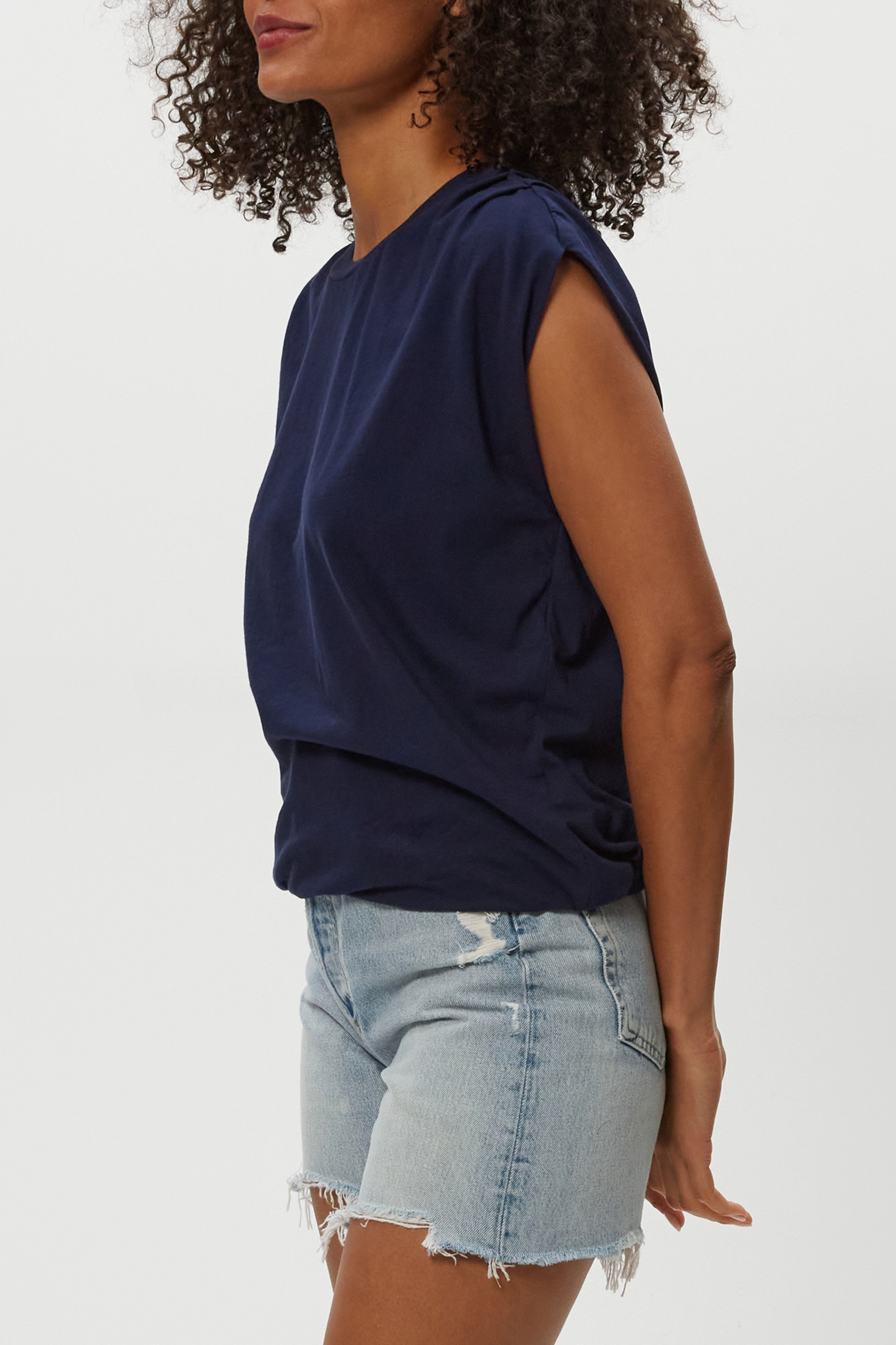 A woman with curly hair wears the Luca Bubble Tee by Michael Stars in navy blue paired with light blue denim shorts with frayed hems, standing against a plain white background.