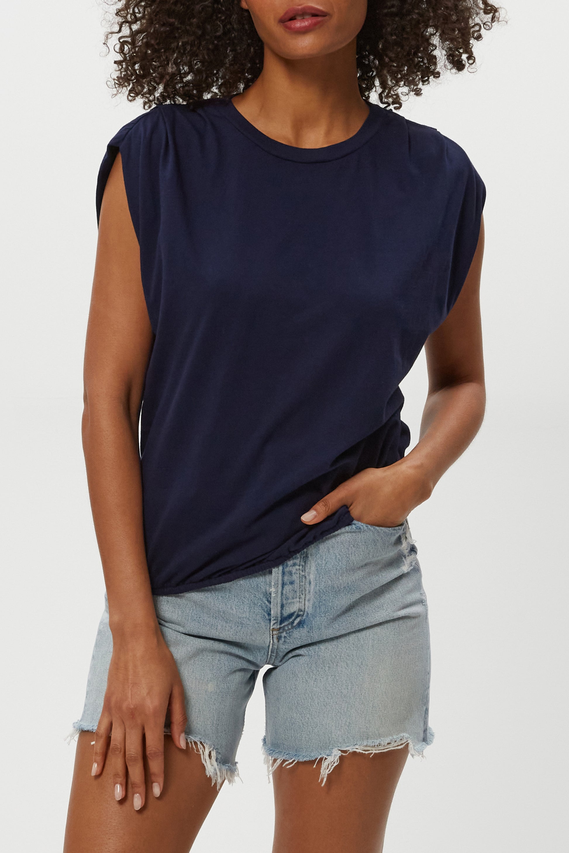 A woman wears the Michael Stars Luca Bubble Tee and light blue denim cutoff shorts, standing with one hand in her pocket against a plain white background.
