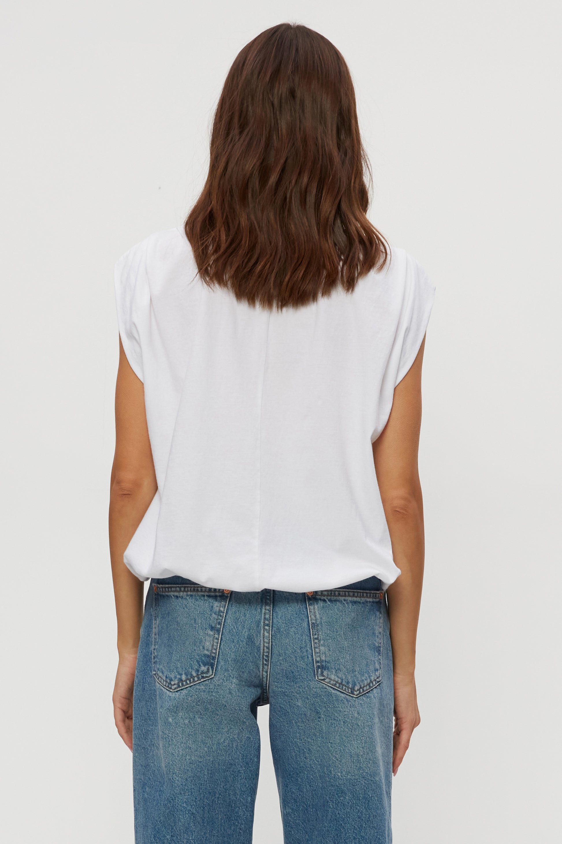 A person with wavy brown hair is seen from the back, wearing the Michael Stars Luca Bubble Tee—a relaxed fit, 100% cotton white sleeveless top—and blue jeans against a plain light background.