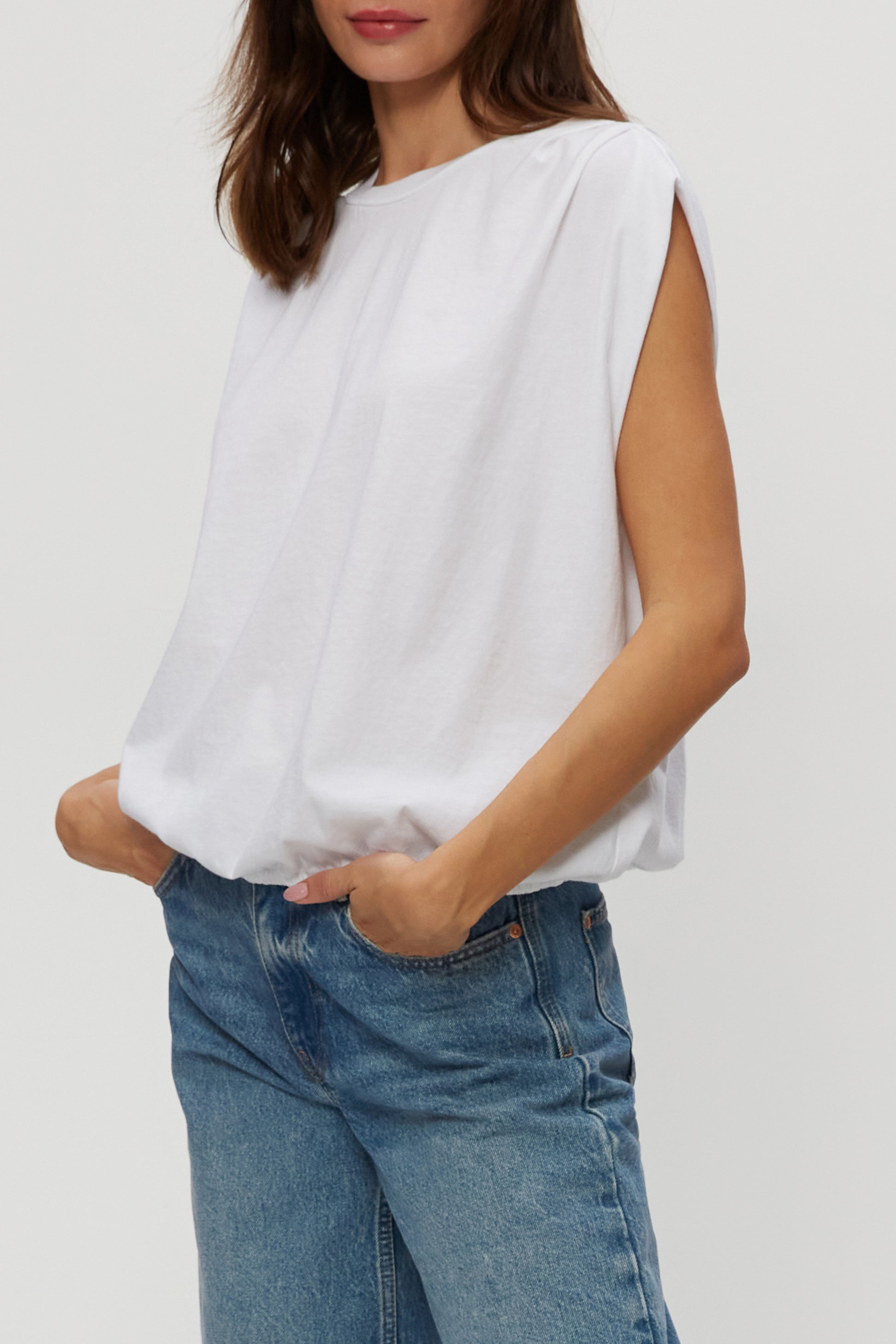 A woman with shoulder-length brown hair models the Michael Stars Luca Bubble Tee—a relaxed fit, sleeveless white top—paired with blue jeans. She stands against a plain light background, hands in pockets. Photo is cropped at shoulders and knees.