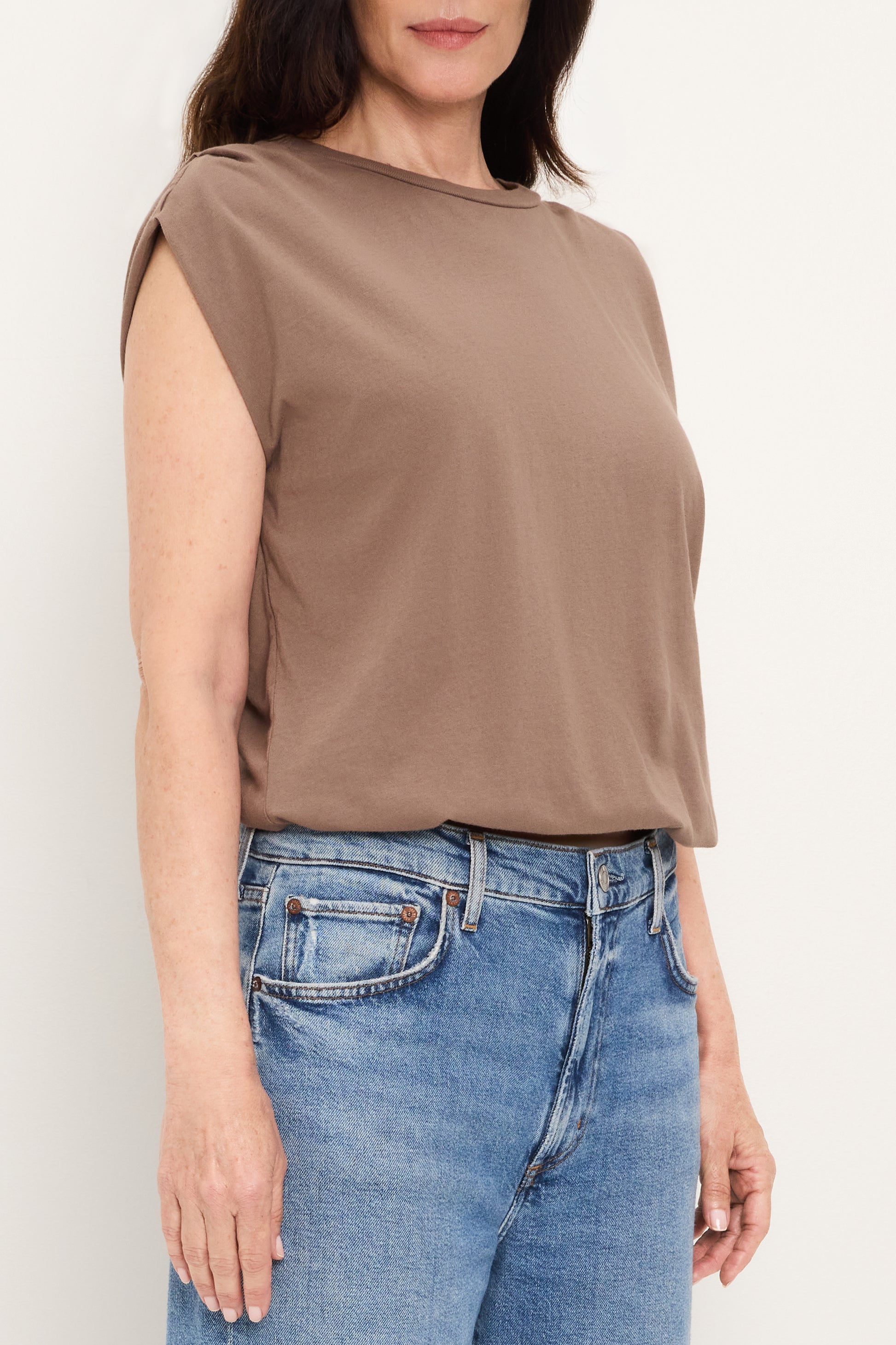 A woman wears the Michael Stars Luca Bubble Tee in Quicksand, a relaxed fit, sleeveless brown top, paired with blue jeans. She stands against a plain light background. The image is cropped from shoulders to hips.