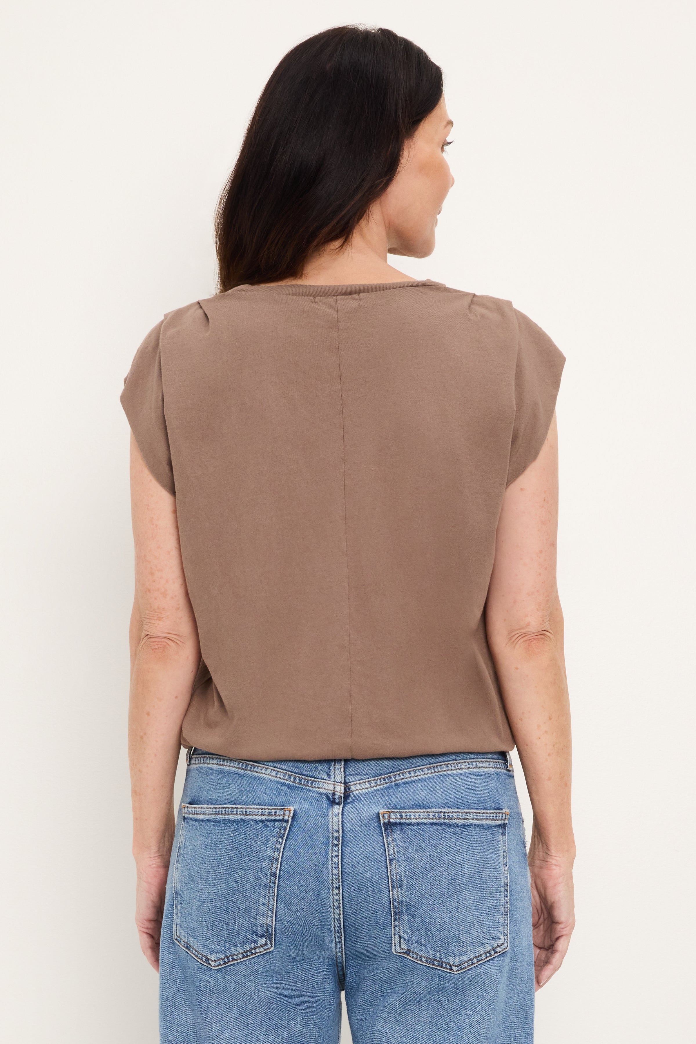 A woman with long dark hair stands facing away, wearing the Michael Stars Luca Bubble Tee in Quicksand and blue jeans against a plain light background.