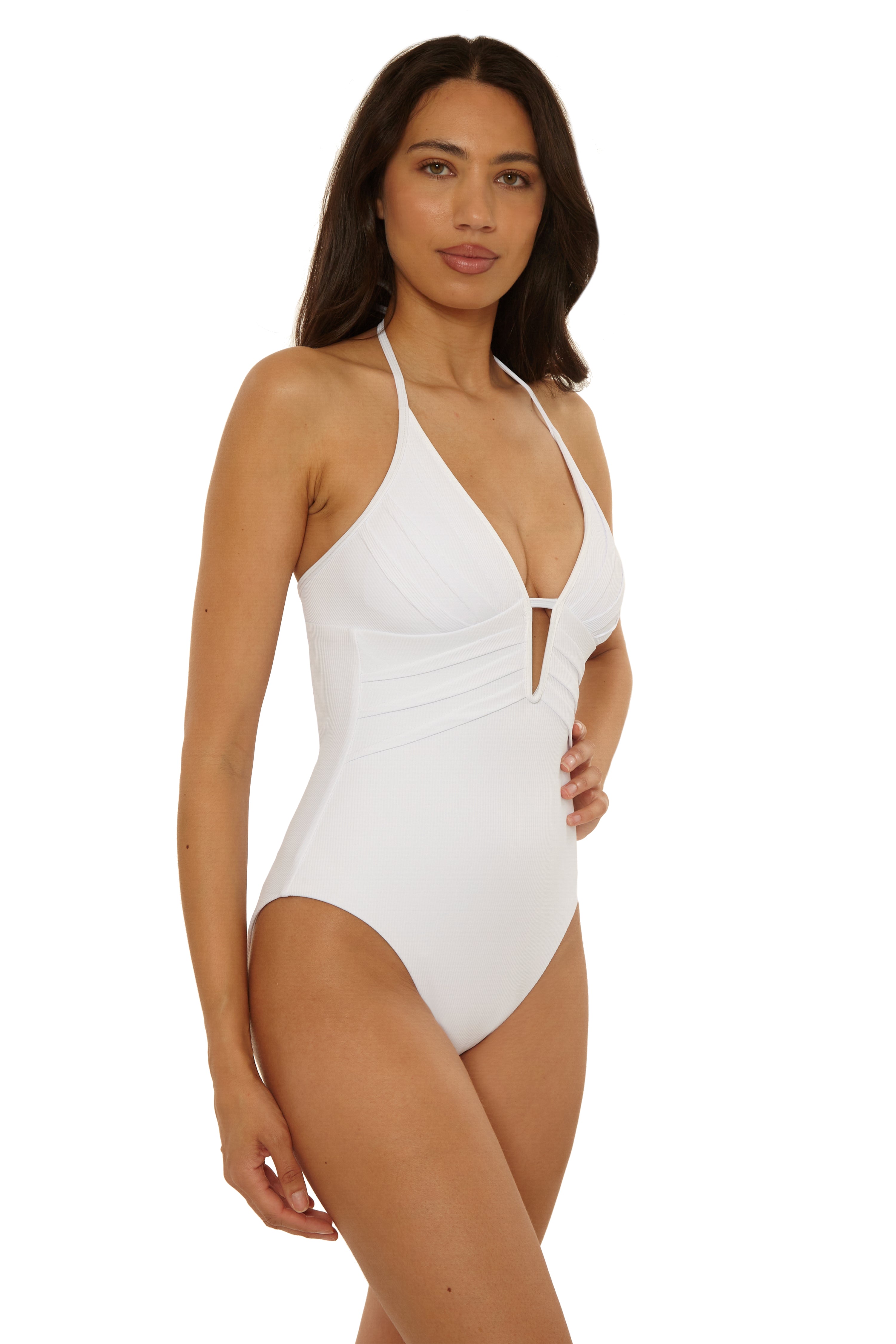 A woman with long dark hair models the Becca Swim Modern Edge One Piece, a white halter-neck swimsuit with a deep V neckline and cutout detail, posing with one hand on her hip against a plain white background.