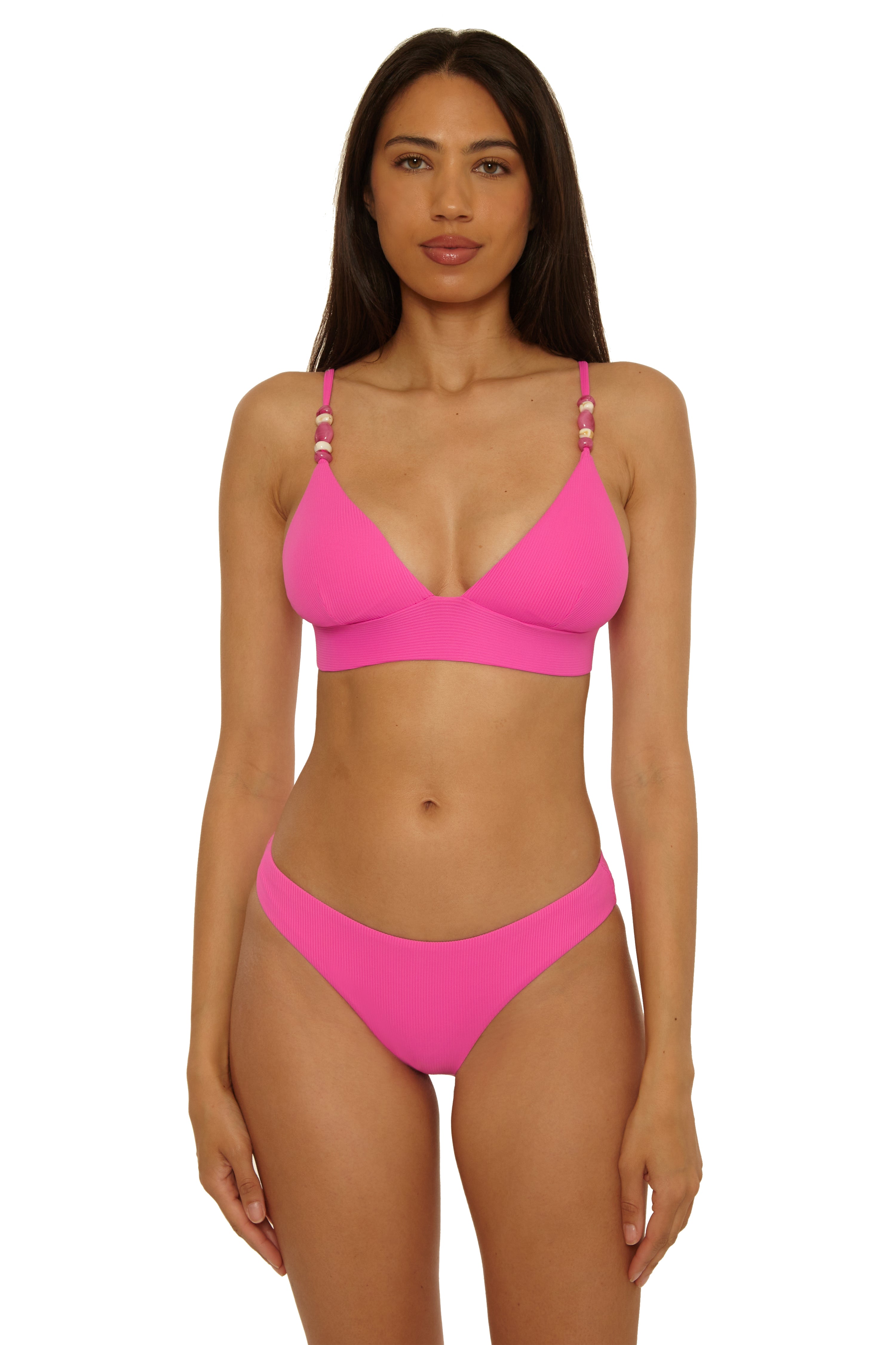 A woman with long brown hair and a neutral expression faces forward in a white background, wearing the Becca Swim Modern Edge Hipster—a bright pink ribbed bikini with bead-detailed straps.