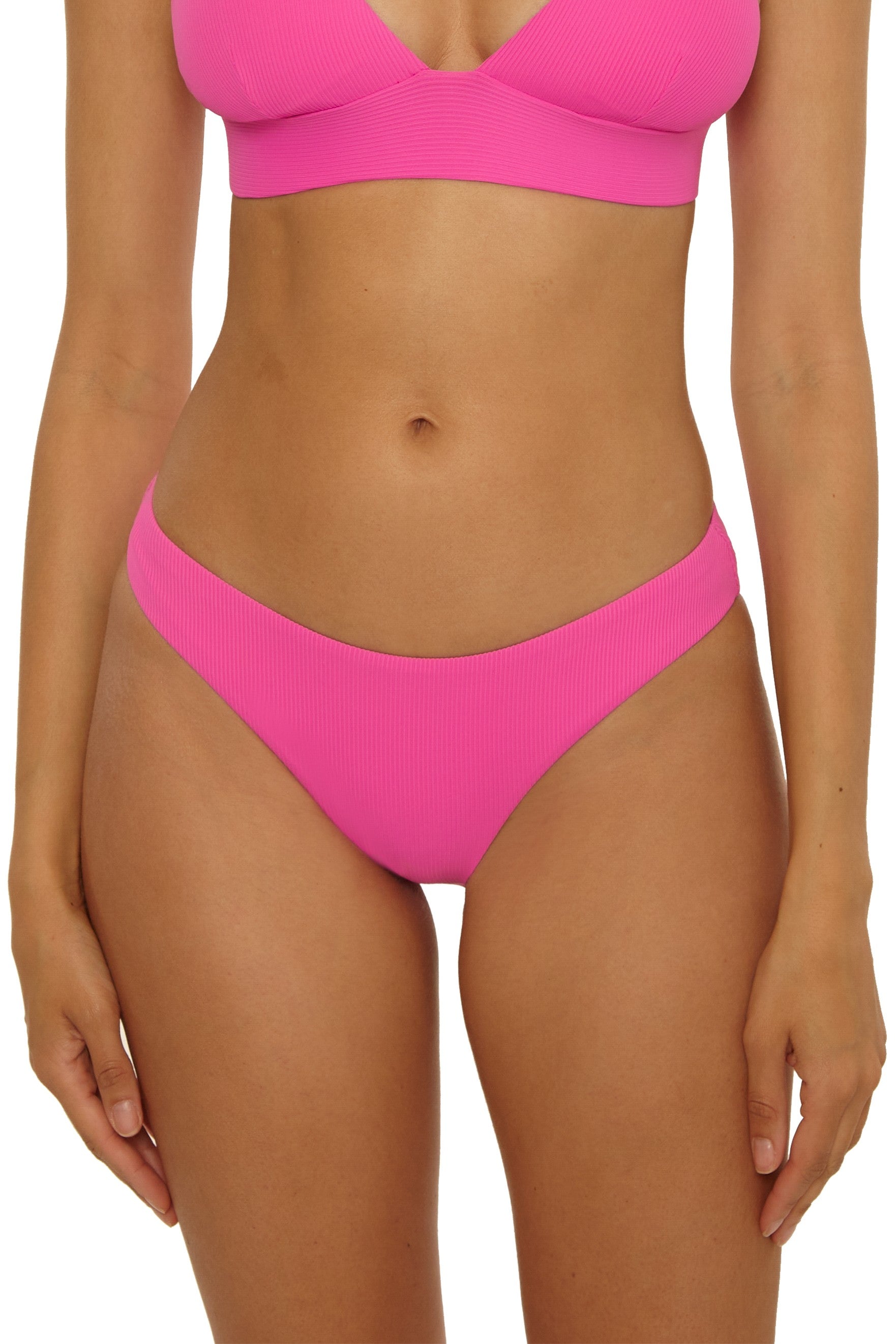 A woman models the Becca Swim Modern Edge Hipster bikini set in bright pink with rib texture, shown from shoulders to upper thighs against a plain background. The hipster bottom is visible and her arms rest naturally by her sides.