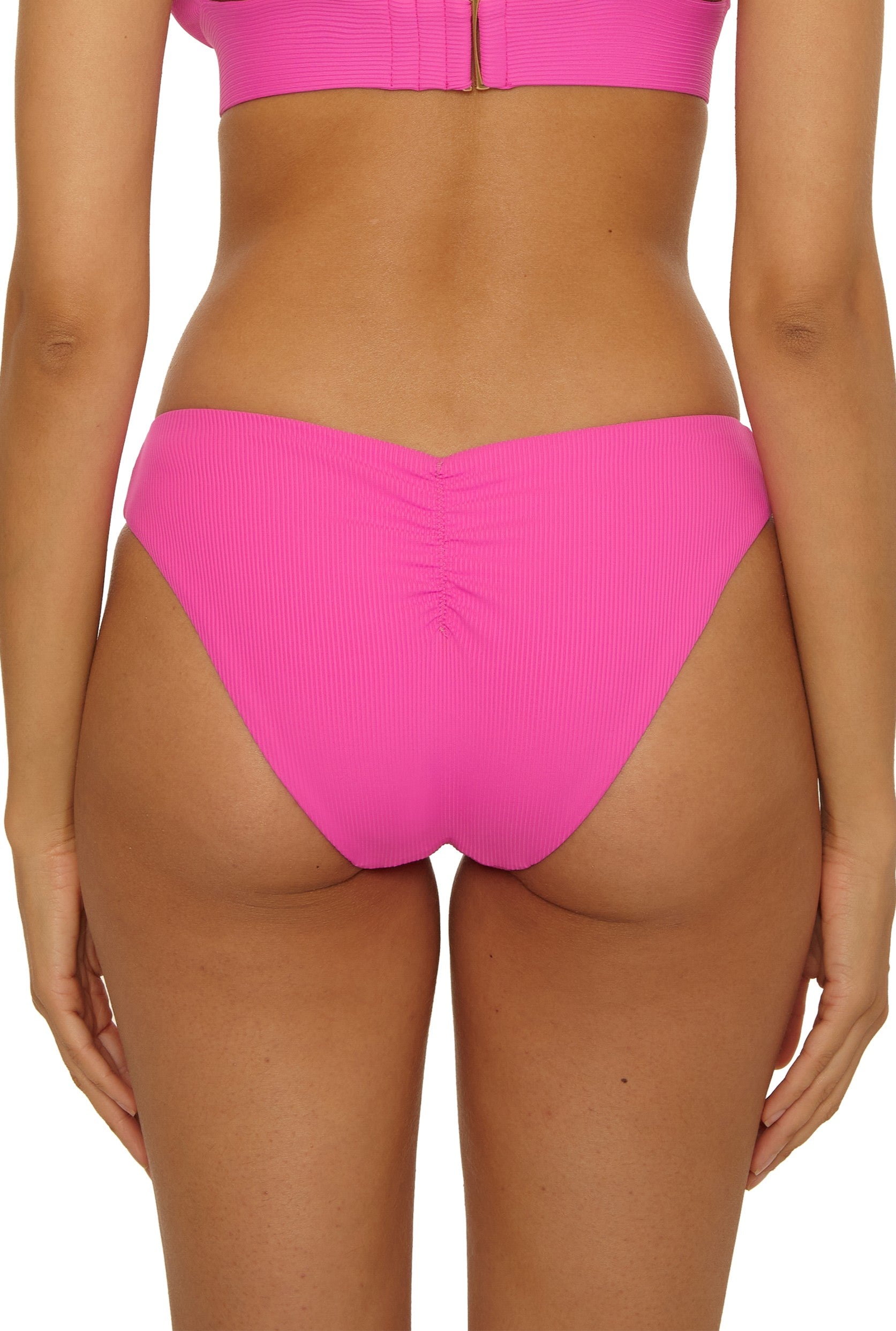 A person wearing Becca Swim's Modern Edge Hipster in bright pink, featuring ribbed texture and ruched back detailing, photographed from behind.